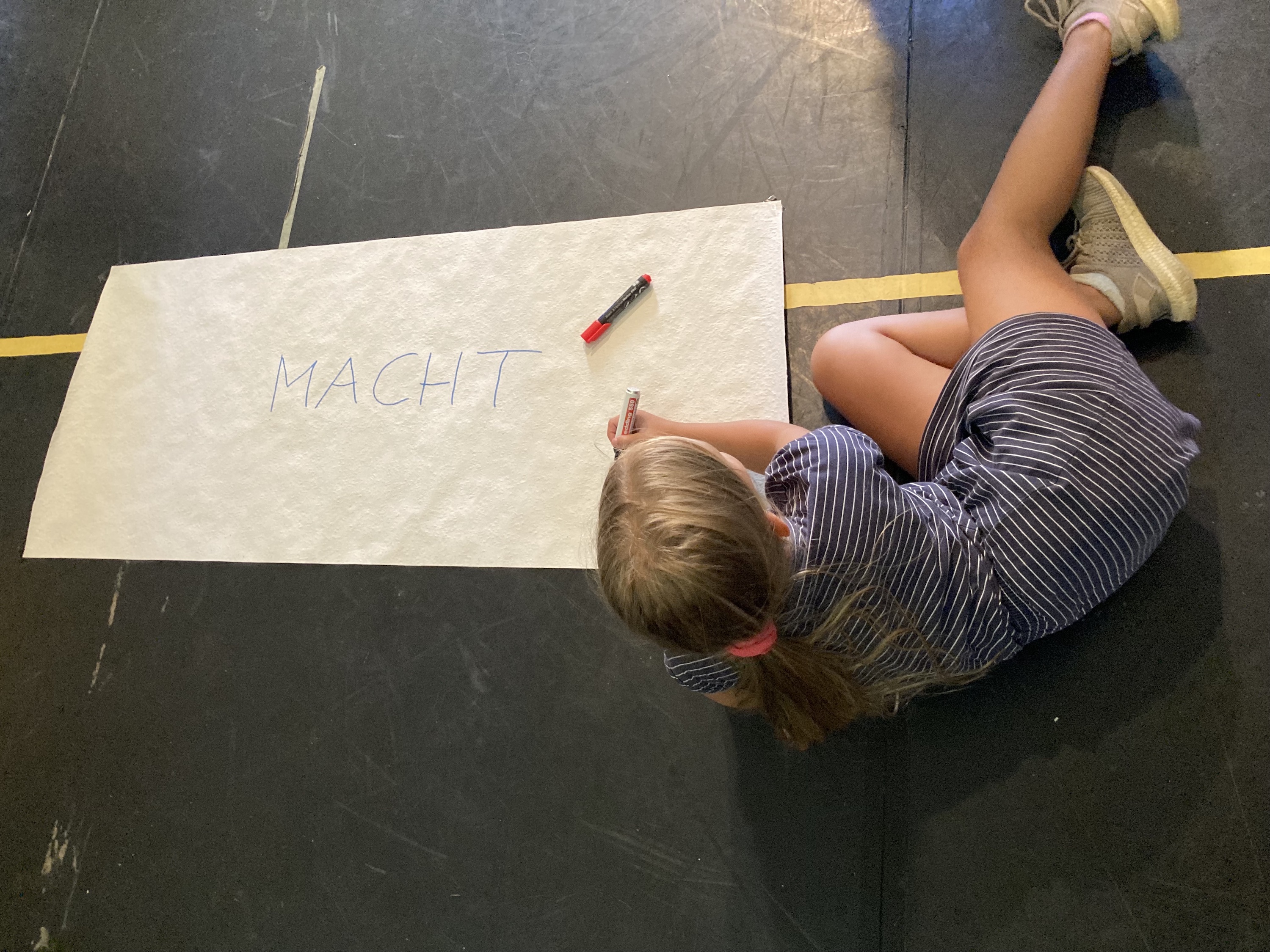 Foto aus Ferienlabor auf Probebühne mit schwarzem Tanzboden. Eine junge Akteurin liegt mit einem gezückten Stift in der Hand über ein Plakat gebeugt. Auf dem Plakat steht das Wort „Macht“ groß in der Mitte. Ein weiterer Stift liegt auf dem Plakat.